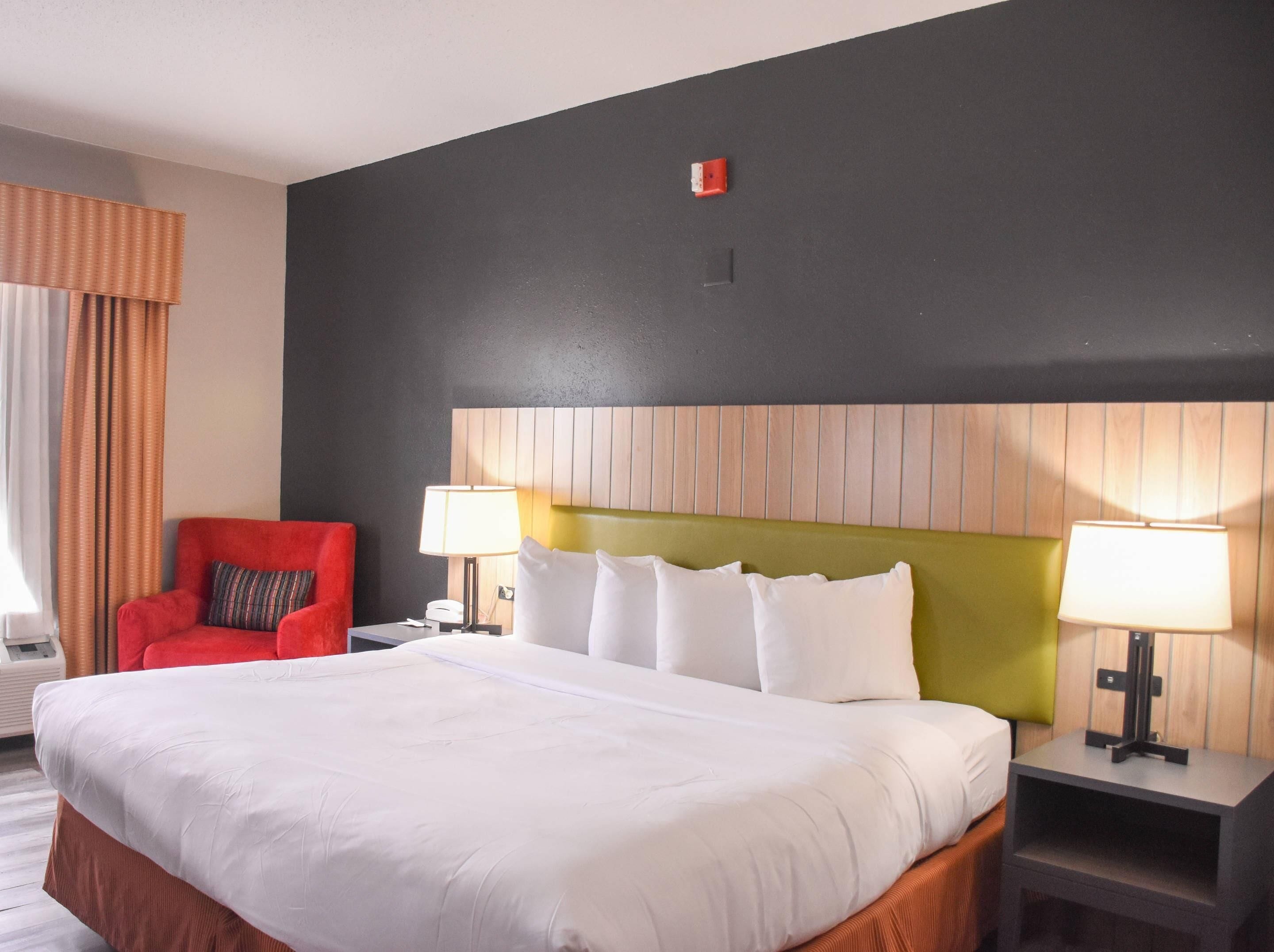 Una camera d'albergo con un letto grande, due comodini con lampade, una poltrona rossa e una testiera imbottita con listelli di legno verticali.