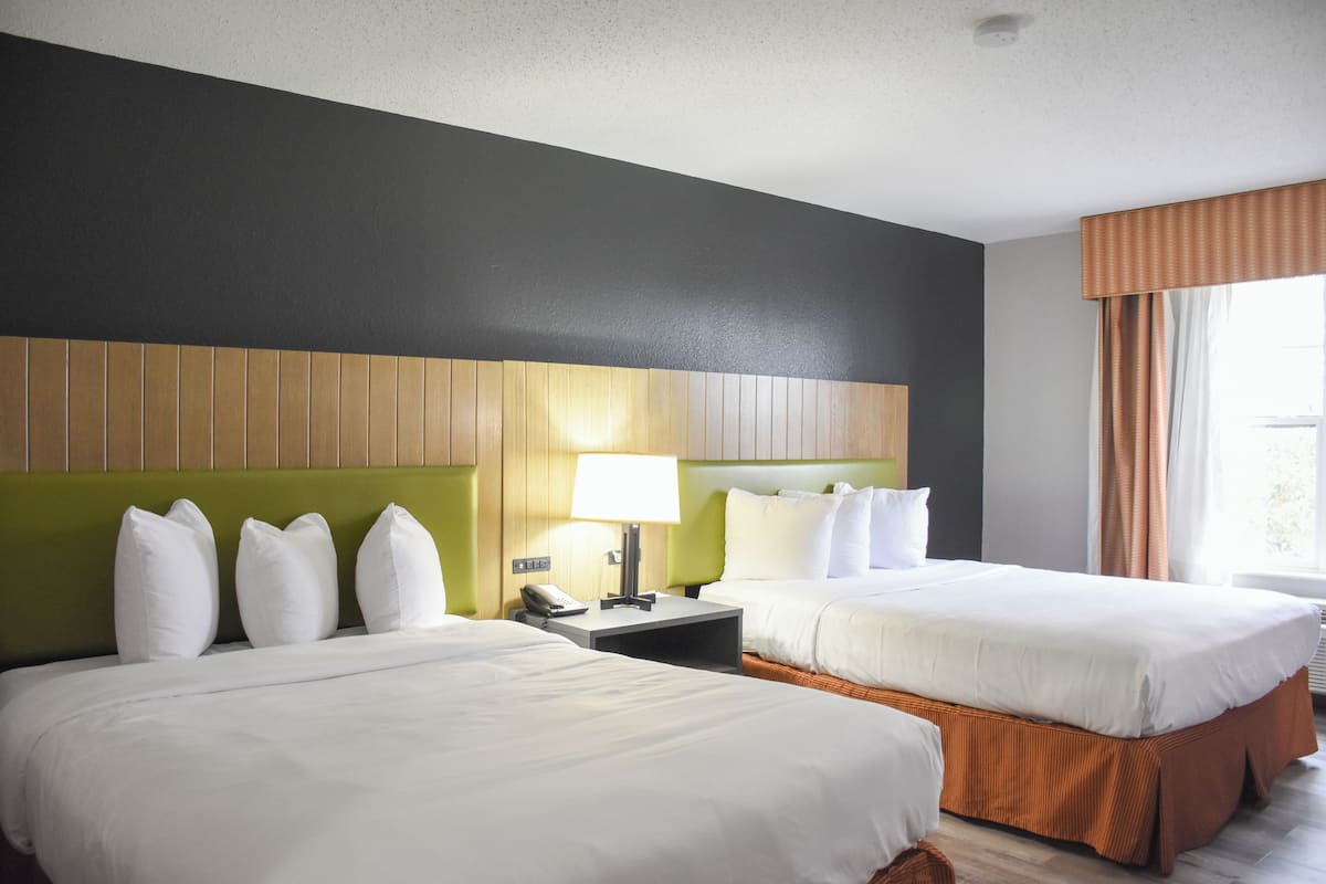 Una habitación de hotel con dos camas, un cabecero verde, una mesita de noche con lámpara y una ventana con cortinas.