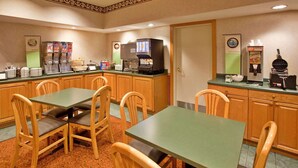A hotel room with a kitchenette, including a coffee maker, a waffle maker, and a refrigerator. There are four wooden chairs and a green table.