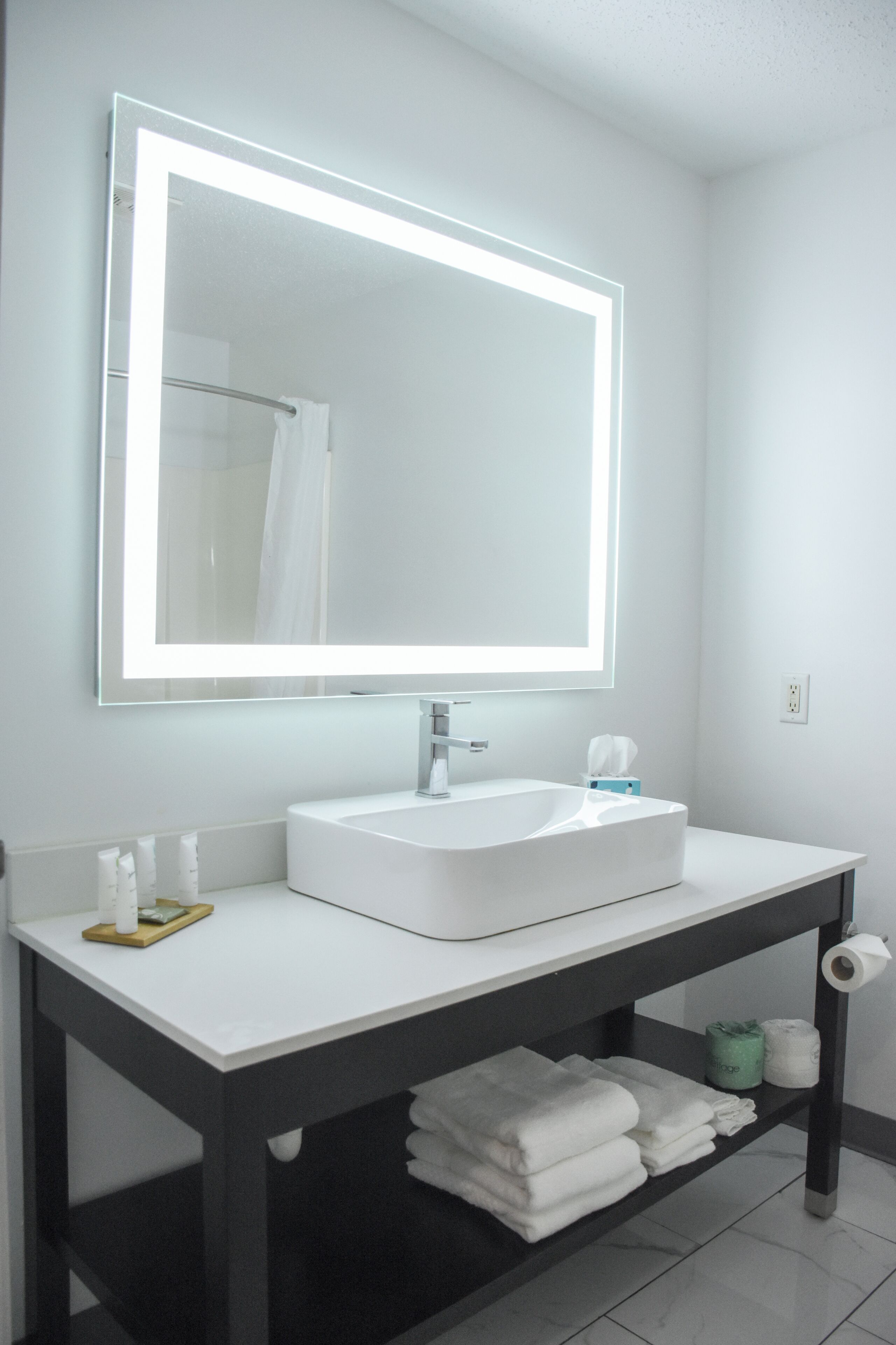 A modern bathroom with a white sink, a large mirror with LED lighting, and neatly folded towels on a shelf.
