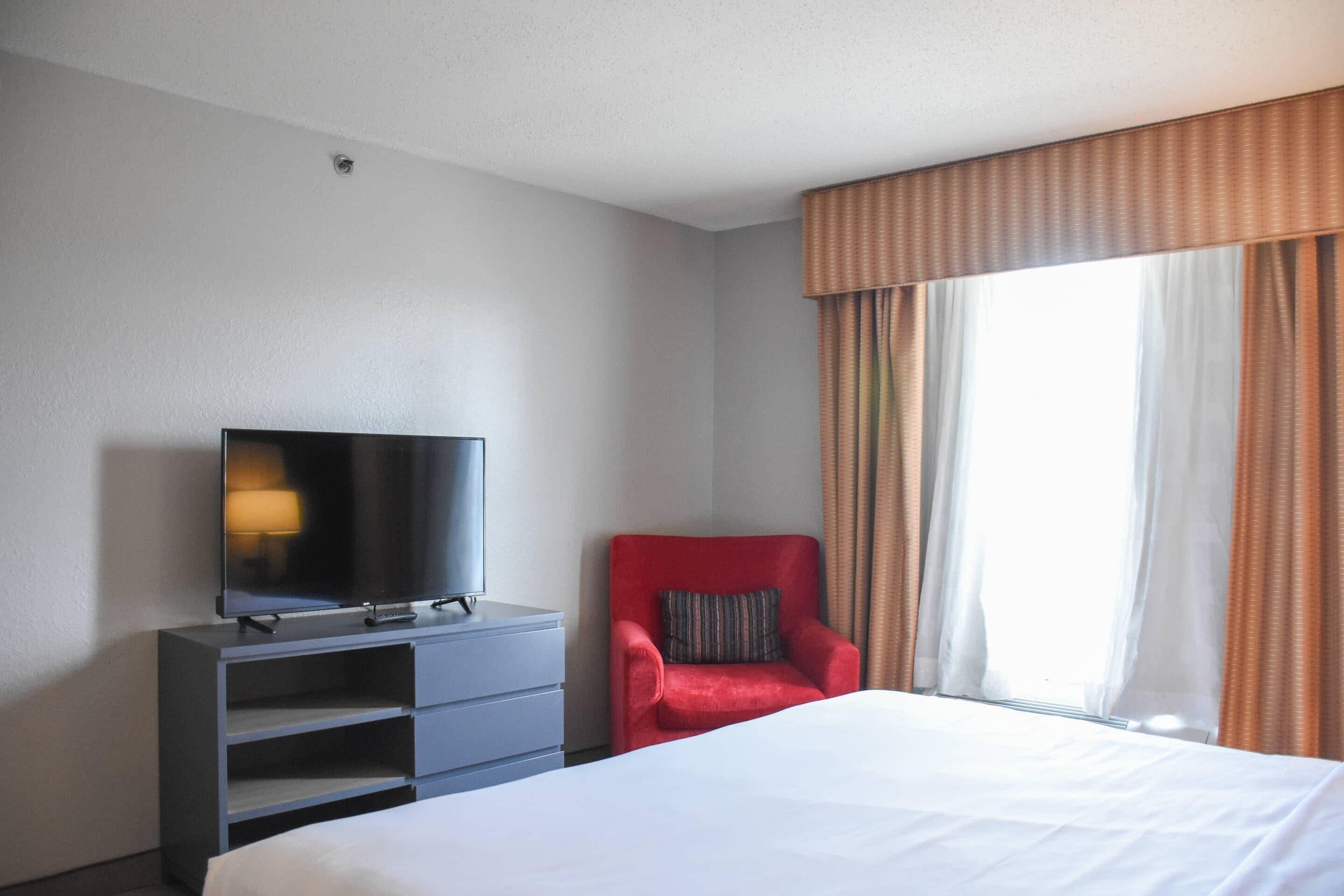 A hotel room with a bed, a television on a dresser, a red armchair, and a window with curtains.