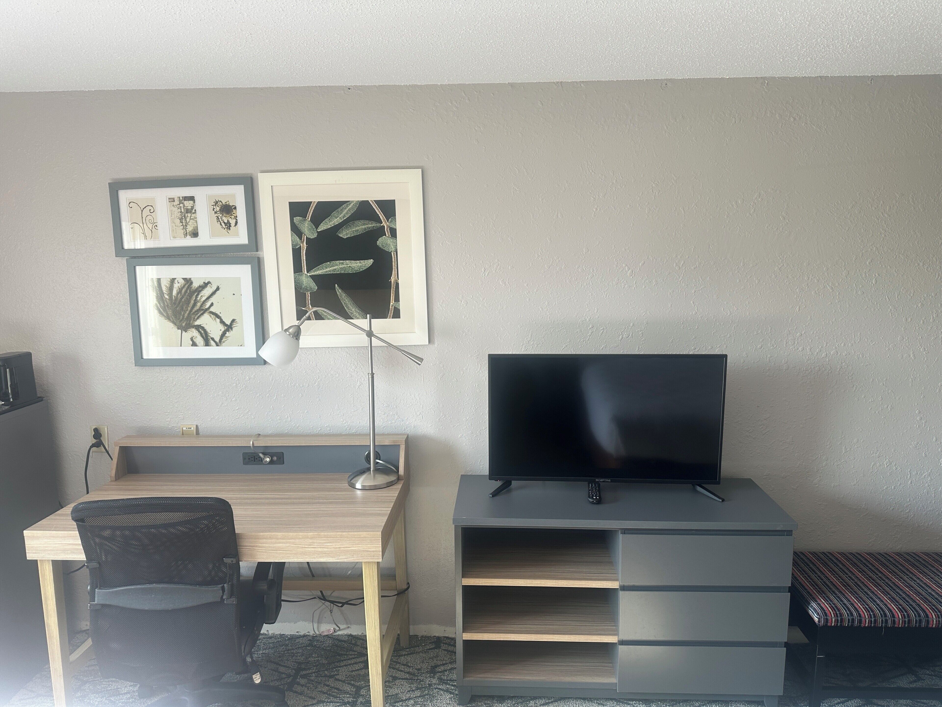 Een kamer met een bureau, stoel, televisie en kunstwerk aan de muur.