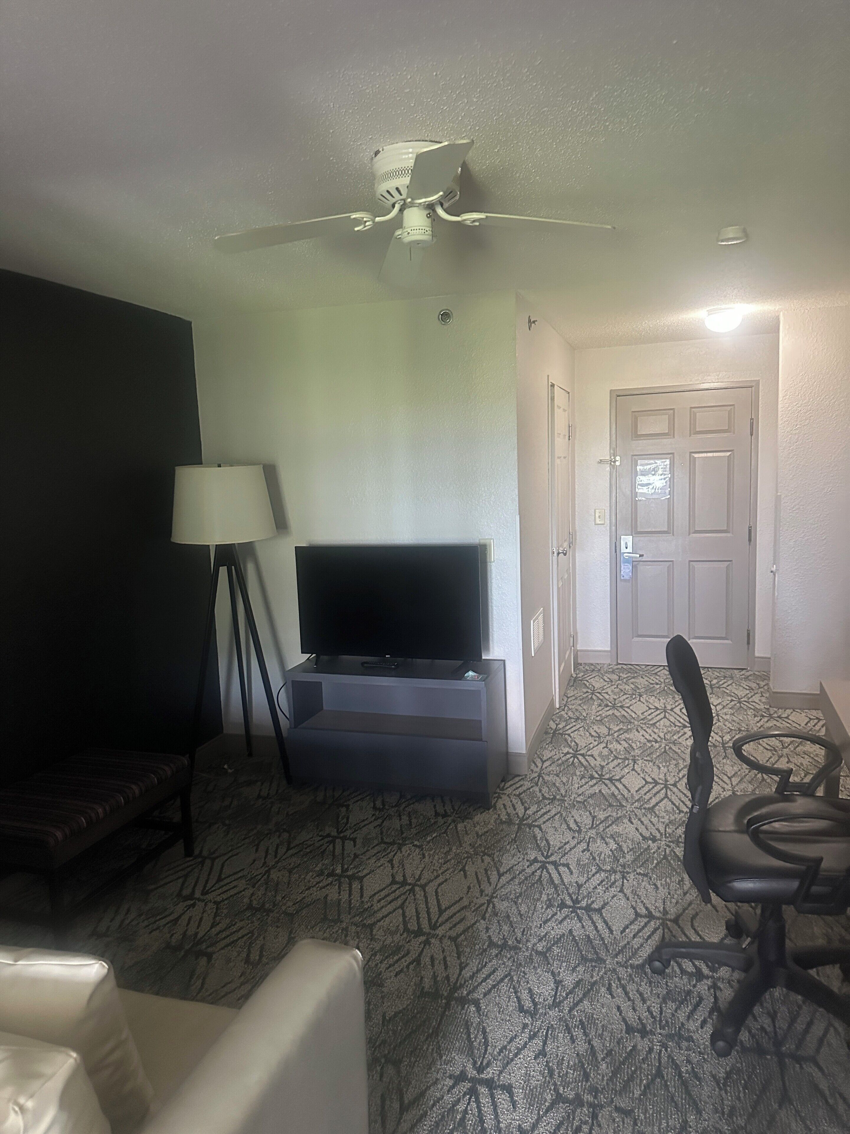 A hotel room with a flat-screen TV, a ceiling fan, a lamp, a sofa, a chair, and a patterned carpet.