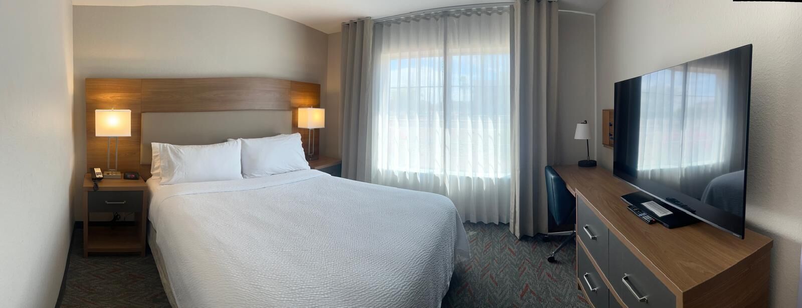 Una habitación de hotel con una cama grande, un escritorio con televisor de pantalla plana, una mesita de noche con lámpara y una ventana con cortinas traslúcidas.