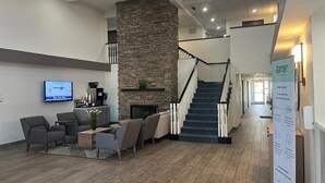 A lobby with a stone fireplace, a TV mounted on the wall, and a staircase leading to another floor. - Garner Hotel Phoenix Northwest – Surprise by IHG (Surprise)