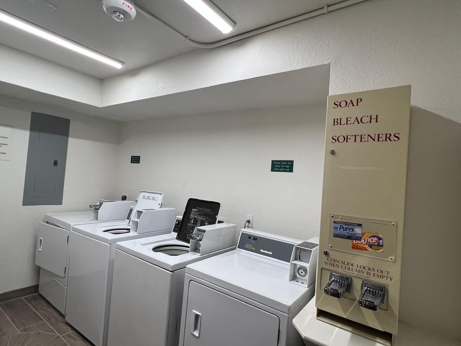 A laundry area with multiple washers and dryers, a soap and bleach softener dispenser, and a coin-operated dryer trap cleaning machine.
