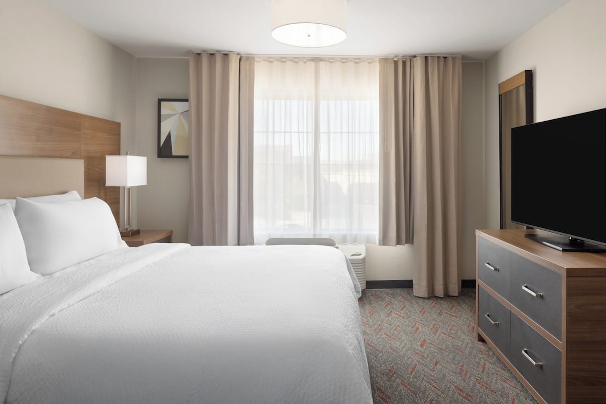 Une chambre d’hôtel avec un grand lit, un téléviseur posé sur une commode en bois, une lampe de chevet et une fenêtre avec des rideaux.