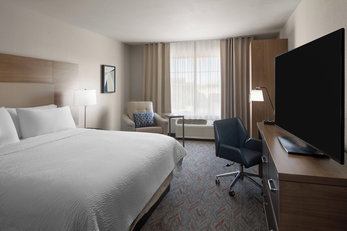 A hotel room with a large bed, a desk with a flat-screen TV, a chair, a lamp, and a window with curtains.