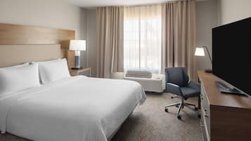 A hotel room with a large bed, a desk with a chair, a TV, a lamp, an air conditioning unit, and a window with curtains.