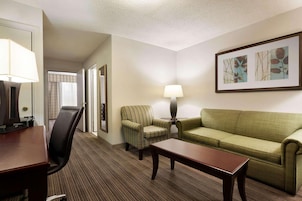 Quarto de hotel com escrivaninha de madeira, sofá verde, poltrona listrada, luminária e um quadro na parede.