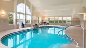 An indoor swimming pool with clear blue water, large windows, and a sunlit room with a table and chairs.
