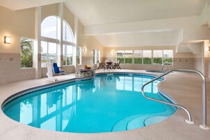 Piscina interior com água azul cristalina, grandes janelas e um ambiente iluminado pelo sol, com mesa e cadeiras.