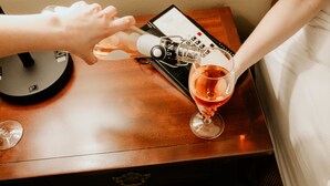 A person pouring a drink into a glass on a wooden table next to a black device with a screen.