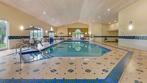 An indoor swimming pool with tiled flooring, a wooden ceiling, and a seating area with chairs and umbrellas.
