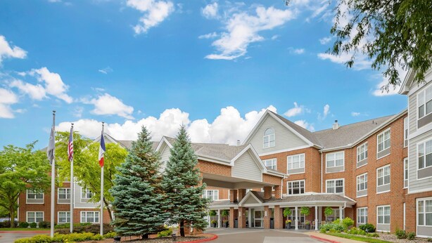 A multi-story brick building with a covered entrance, flagpoles, and a landscaped area with trees.