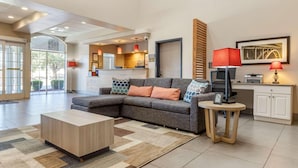 A modern living room with a grey sectional sofa, a wooden coffee table, and a small round table with a lamp. There is a kitchen area in the background with a red lamp and a framed picture on the wall. - Country Inn & Suites by Radisson, Lumberton, NC (Lumberton)