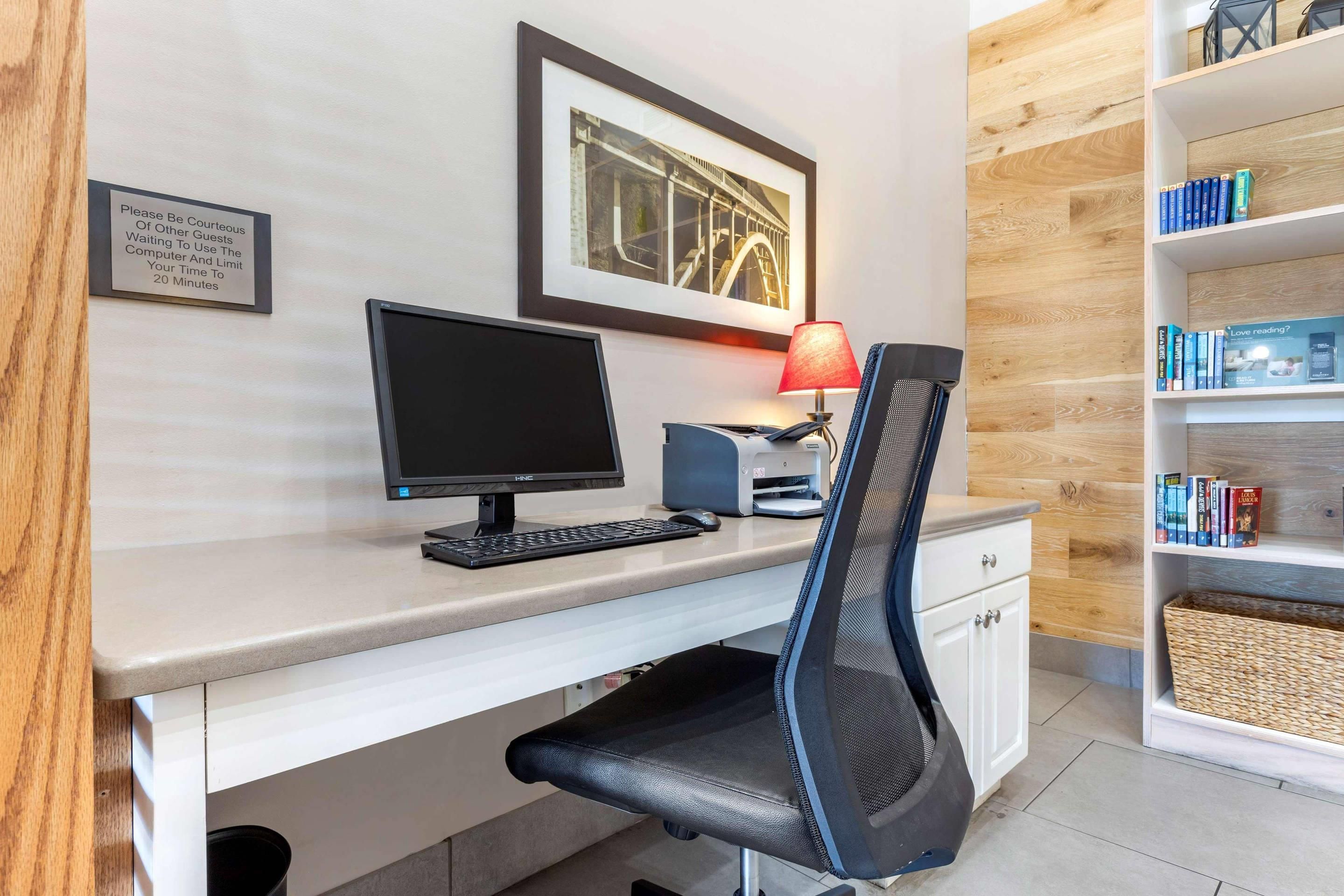 A hotel room with a desk, computer, chair, and bookshelf.
