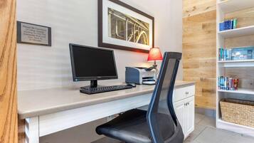 A hotel room with a desk, computer, chair, and bookshelf.