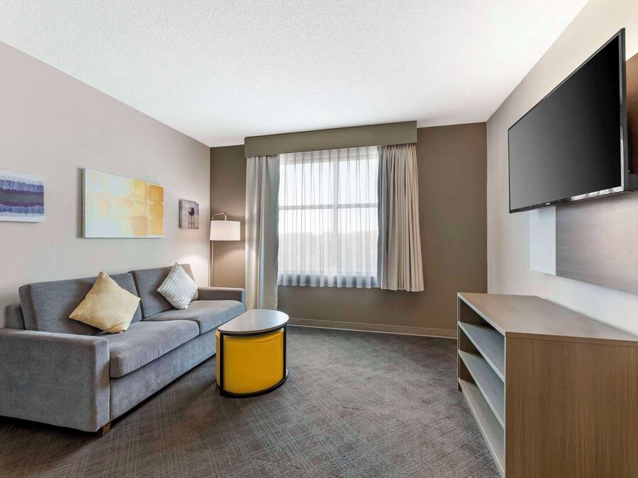 A modern hotel room with a grey sofa, a yellow side table, a flat-screen TV mounted on the wall, and a large window with sheer curtains.