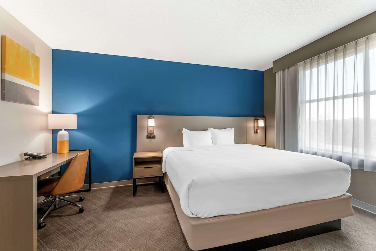 A hotel room with a large bed, a desk with a chair, a nightstand, and a wall-mounted lamp.
