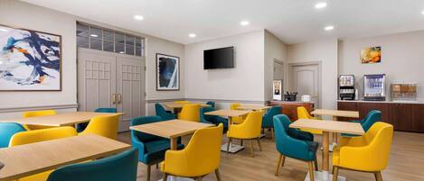 Un espace repas moderne avec des chaises jaunes et turquoise, des tables en bois et un téléviseur à écran plat fixé au mur.