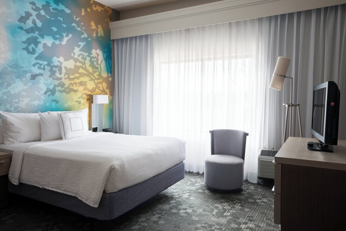 A hotel room with a large bed, a desk with a television, a chair, and a window with curtains.