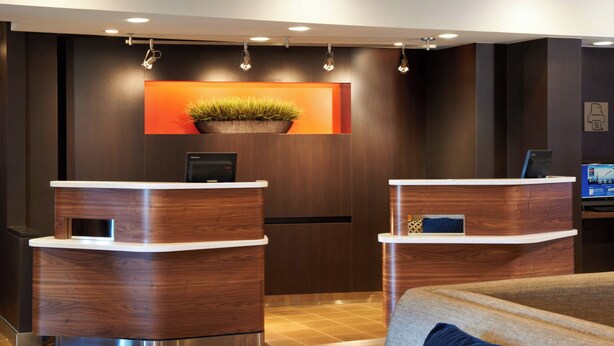 A reception area with two wooden reception desks, a potted plant, and a television screen. - Courtyard by Marriott Toledo Maumee/Arrowhead (Maumee)
