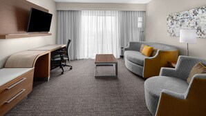 Une chambre d’hôtel moderne équipée d’un téléviseur à écran plat fixé au mur, d’un bureau avec une chaise, d’un canapé gris avec des coussins jaunes et d’une lampe.
