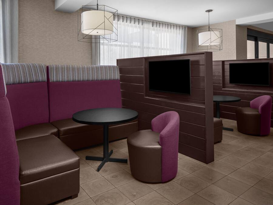 A modern hotel room with a sectional sofa, a round table, a small stool, and a TV mounted on a wooden panel wall.