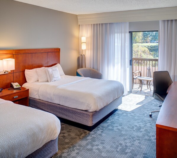 A hotel room with a large bed, a desk with a phone, a chair, and a balcony with a view of trees.