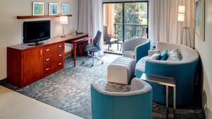 Een moderne hotelkamer met een houten bureau, een flatscreen televisie, een comfortabele zithoek met een bank en voetenbankje, en een groot raam met uitzicht op de omgeving.