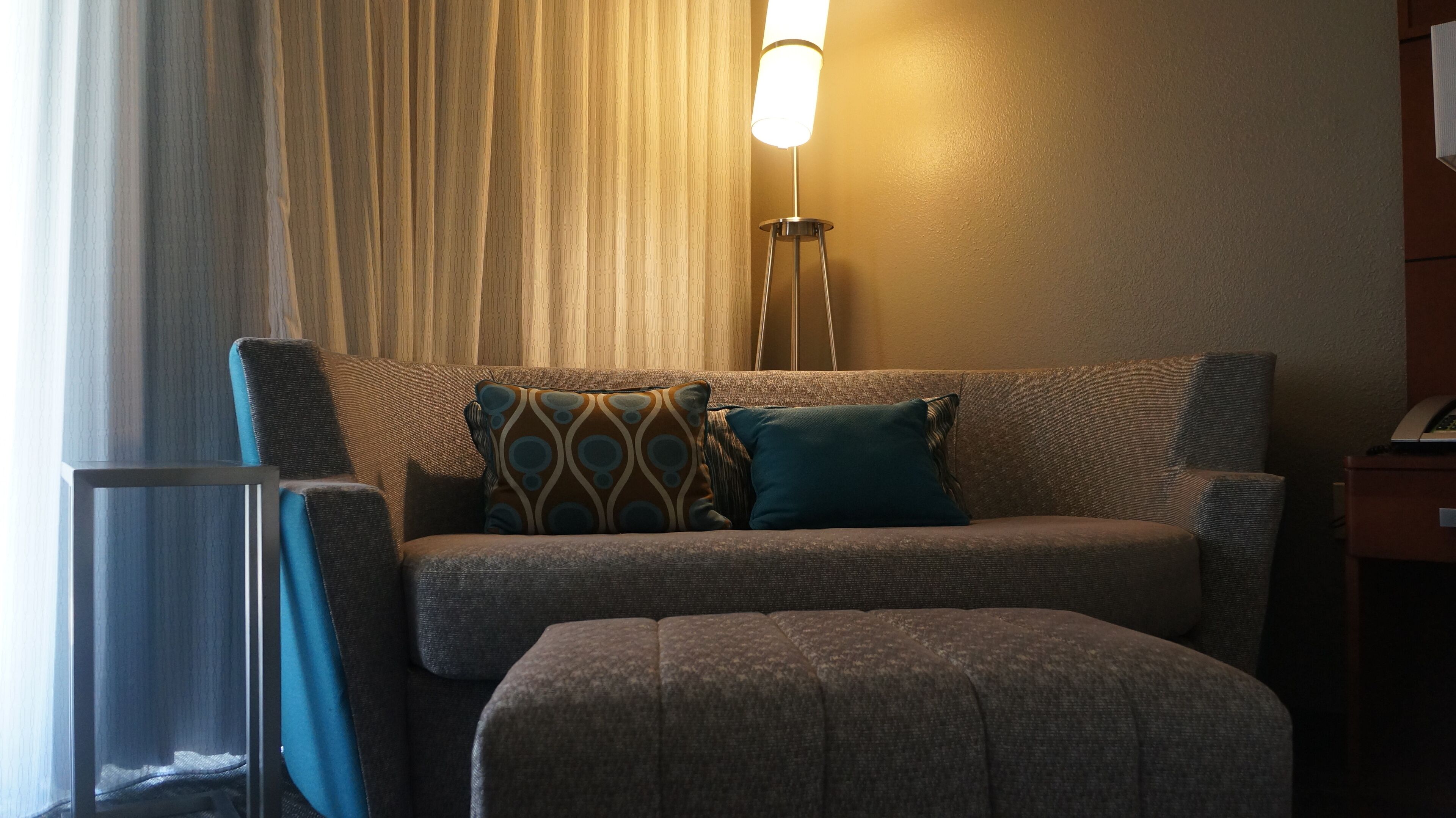 A hotel room with a sofa, two pillows, a standing lamp, and a small table.