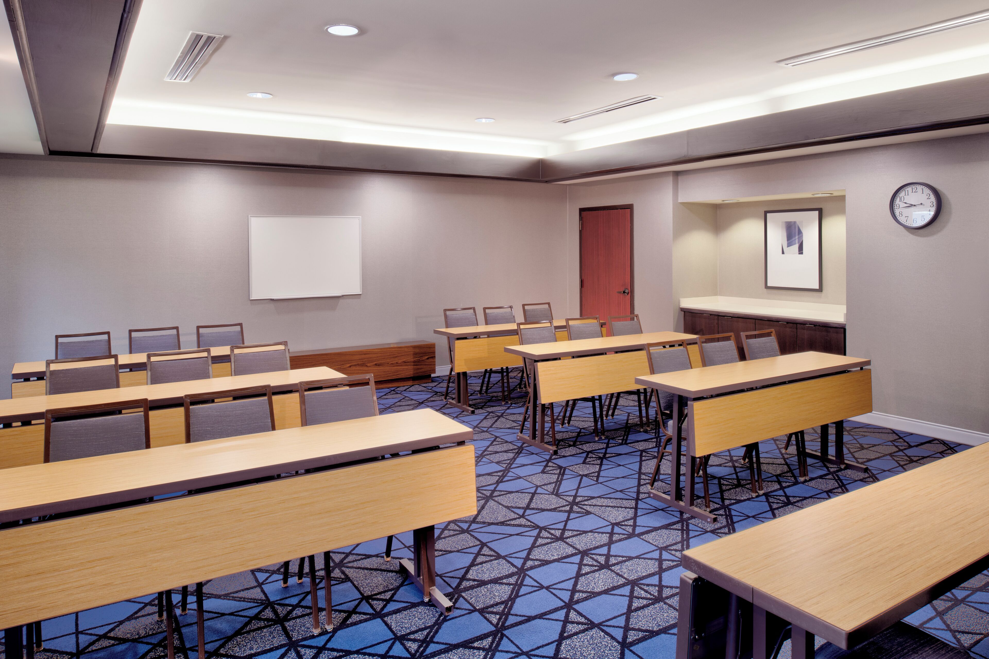 Uma sala de conferências com várias mesas e cadeiras, um relógio na parede e uma porta.