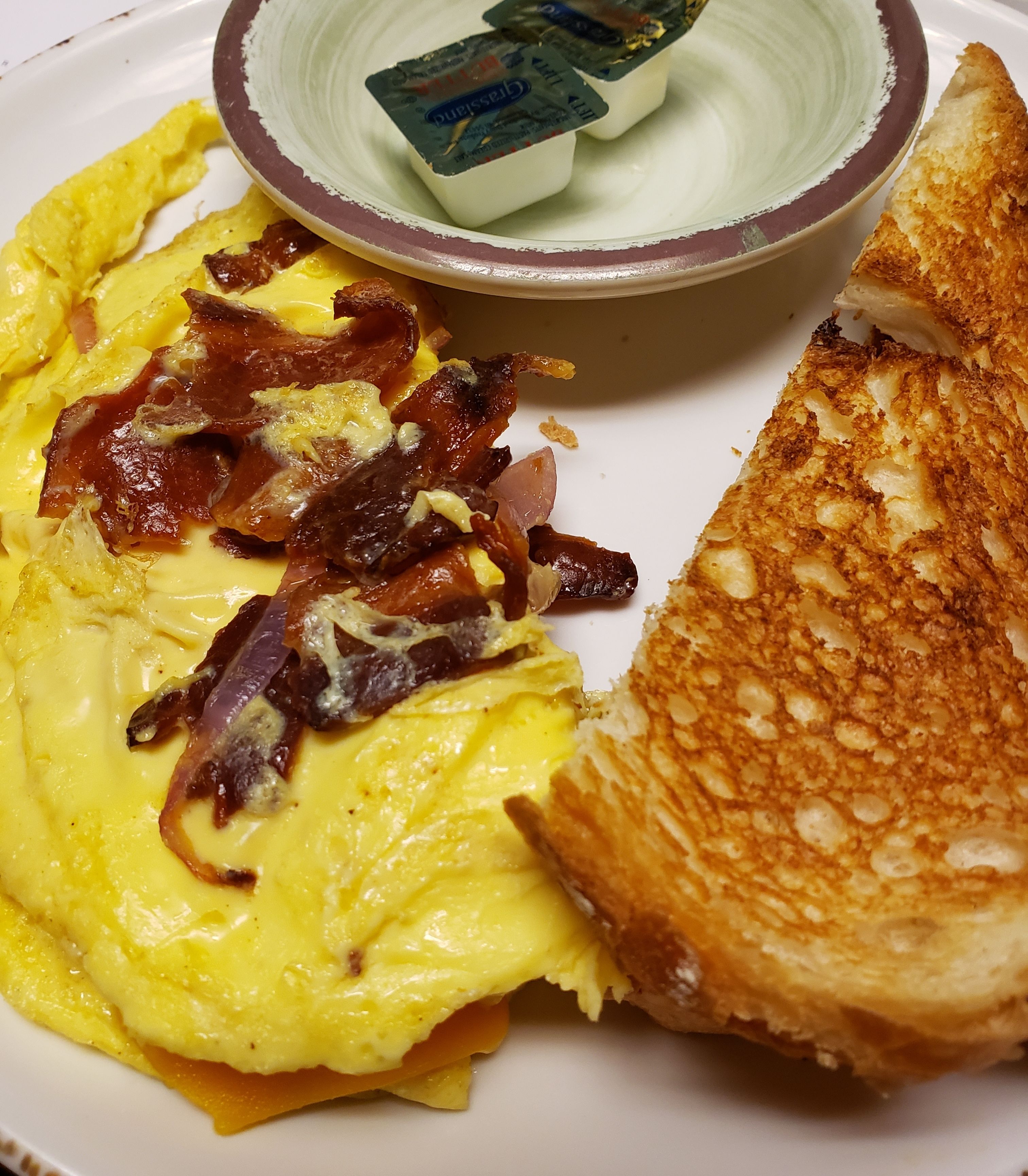 A plate with scrambled eggs, bacon, toast, and a small bowl of yogurt.