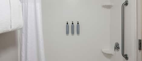 Un baño con una bañera blanca, ducha y barra de apoyo. Hay tres botellas de loción en la pared.