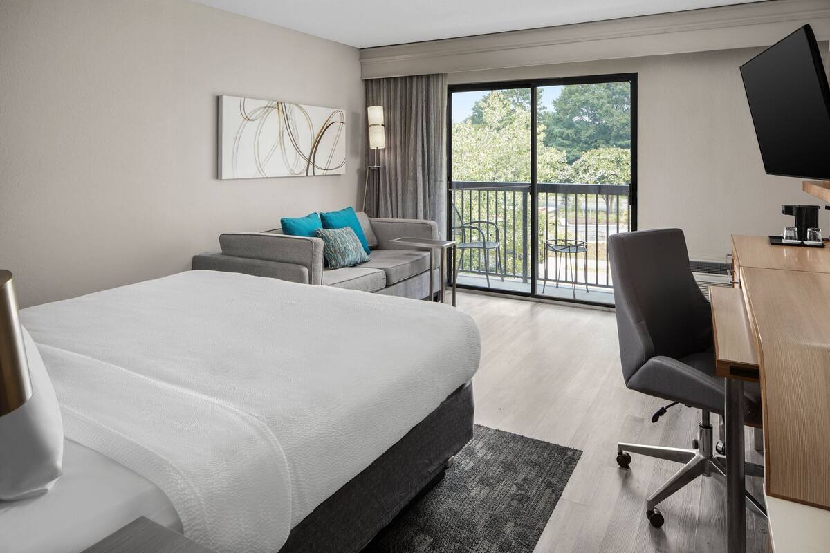 A hotel room with a large bed, a desk, a chair, a TV, and a balcony with a view of trees.