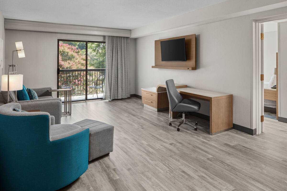 A modern hotel room with a grey sofa, a teal armchair, a wooden desk, and a flat-screen TV mounted on the wall.