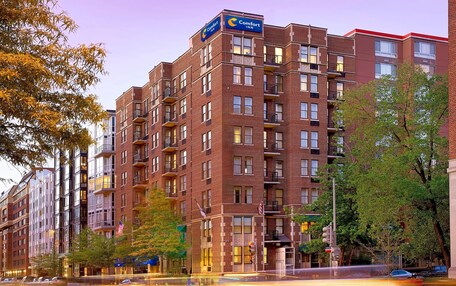 A multi-story brick building with a Comfort Inn sign, featuring balconies, large windows, and a street view with cars and trees.