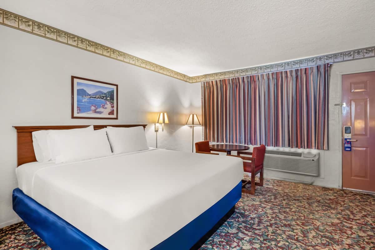 Une chambre d’hôtel avec un grand lit, une petite table avec deux chaises, un tableau au mur et une fenêtre avec des rideaux.