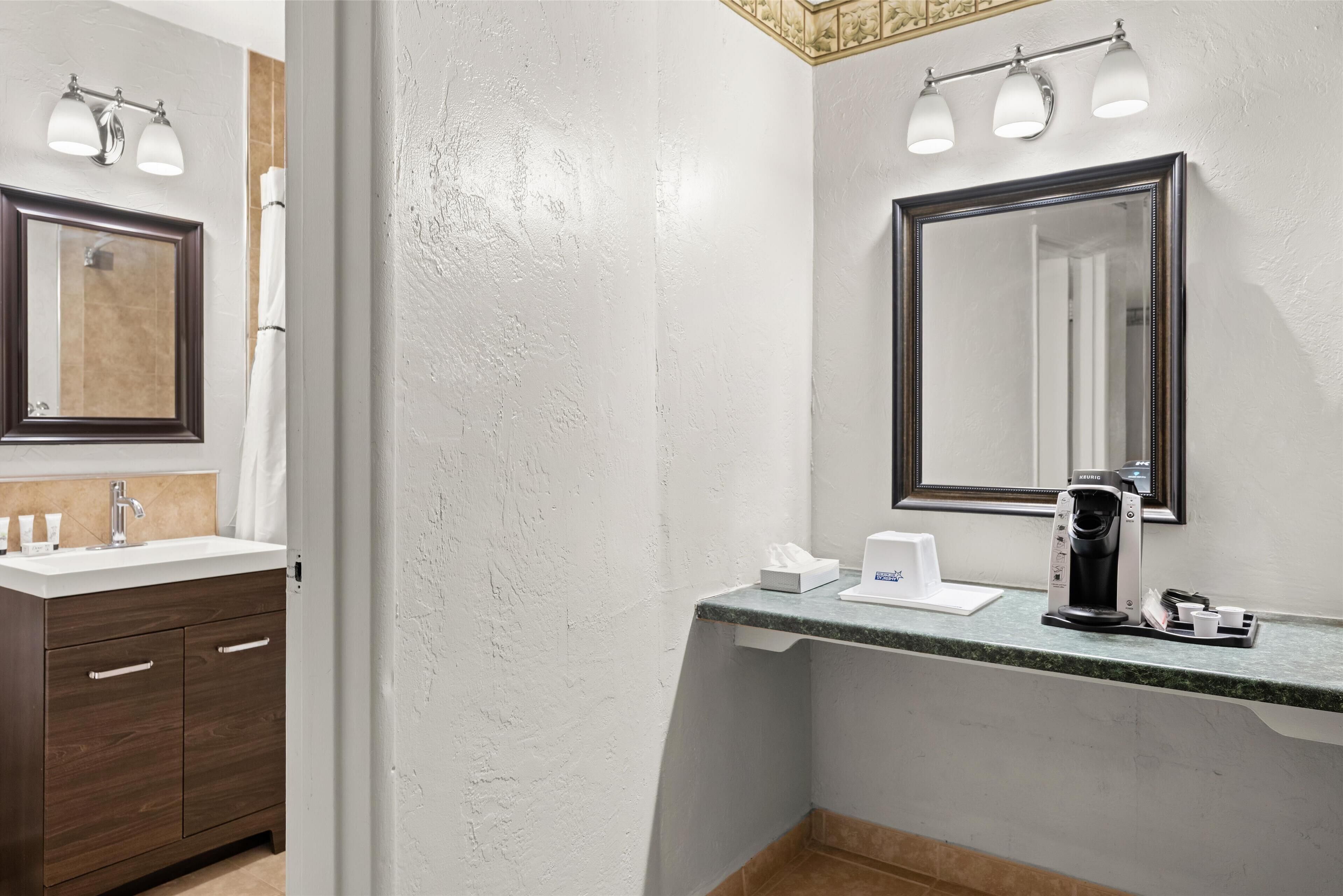 A bathroom with a white sink, dark wooden vanity, and a large mirror. There are three wall-mounted lights above the mirror and a coffee machine on the countertop.