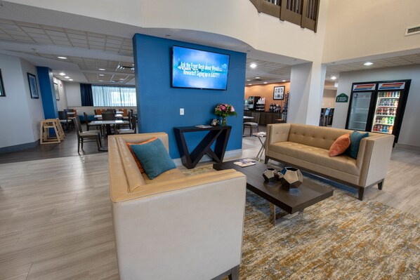 A modern hotel lobby with a large TV screen, comfortable seating, a coffee table, and a refrigerator. - Wingate by Wyndham Indianapolis Airport Plainfield (Plainfield)