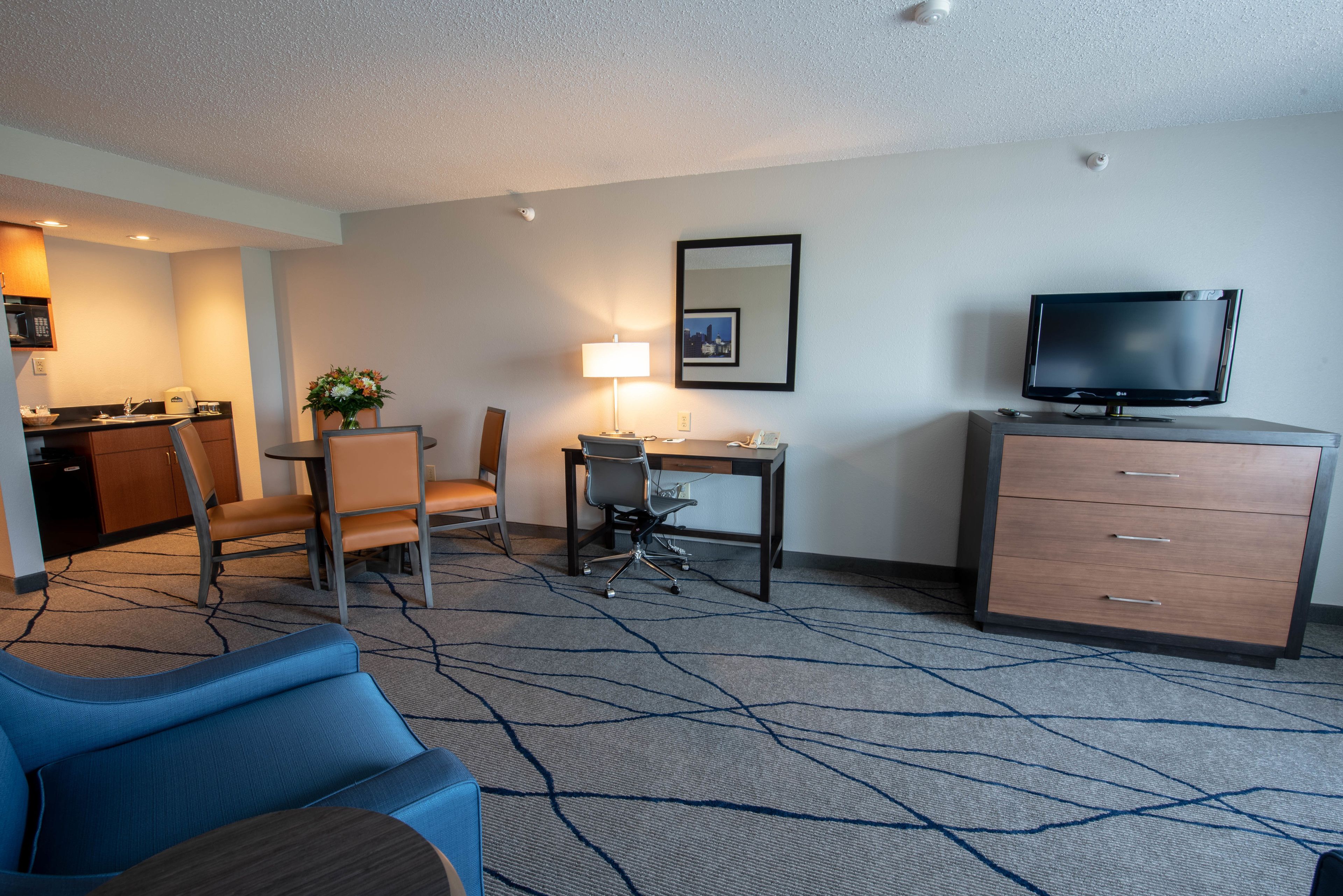 Ein Hotelzimmer mit einer Küchenzeile, einem Essbereich mit Tisch und Stühlen, einem Schreibtisch mit Lampe, einem Fernseher und einem großen, blauen Sofa.