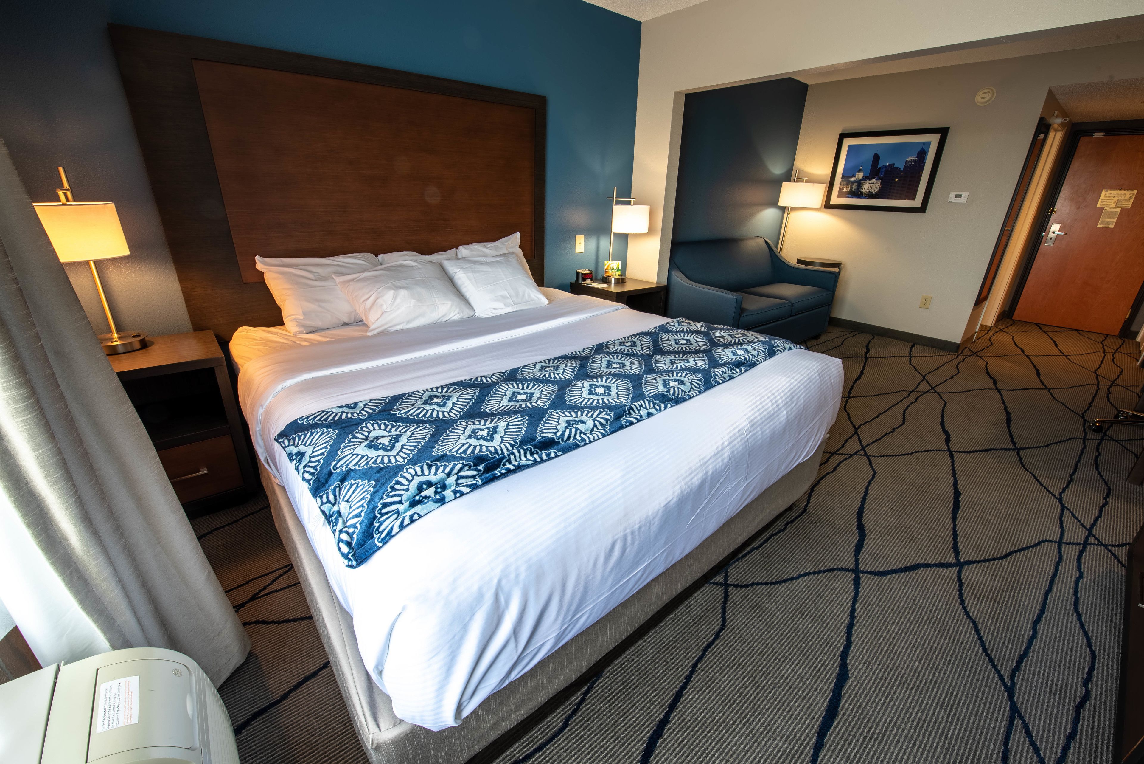 Habitación de hotel con una cama grande, una mesita de noche con lámpara, una silla e imagen enmarcadas en la pared.
