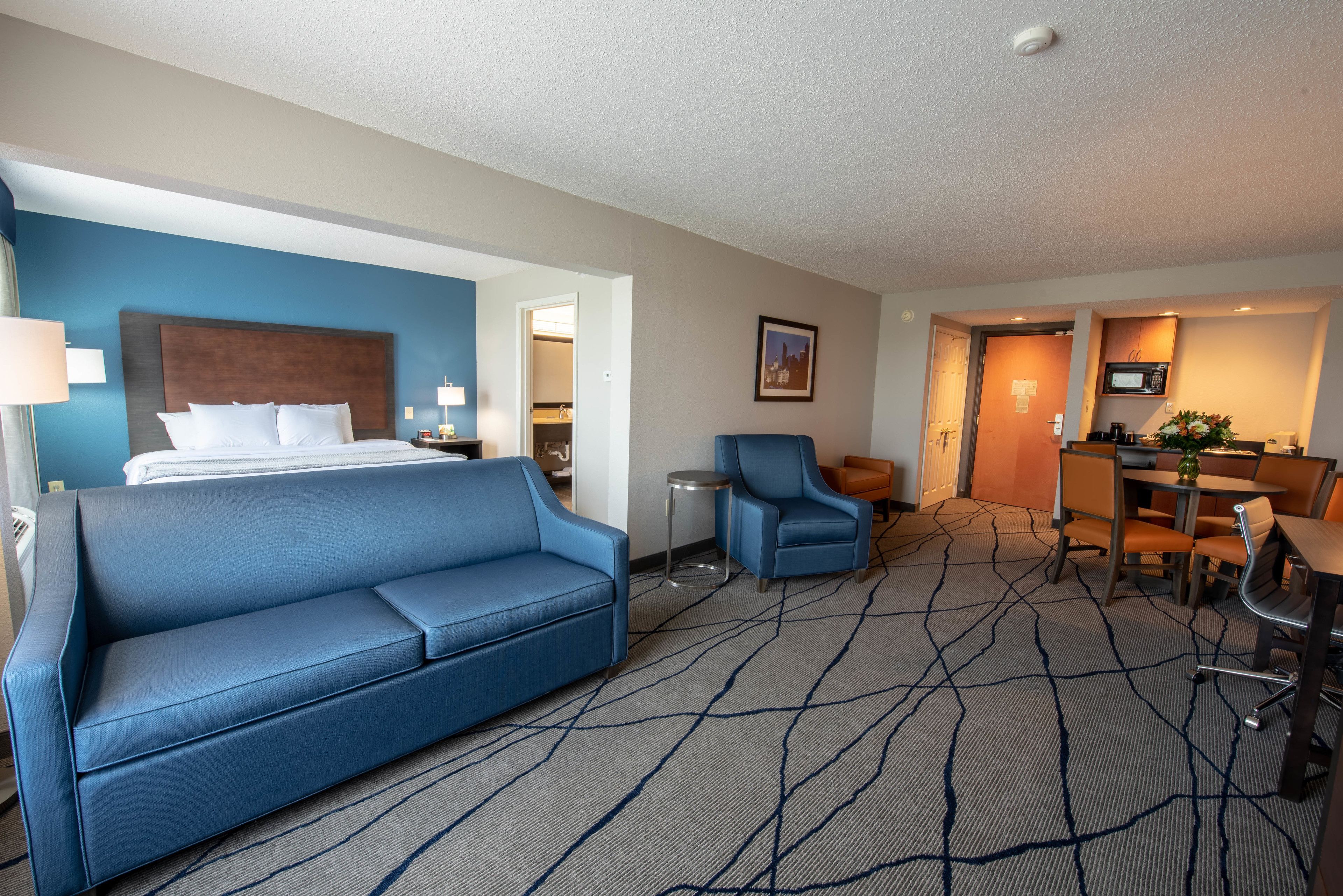 A hotel room with a bed, a blue sofa, a small table, a chair, a microwave, and a dining area with a table and chairs.