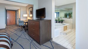 Habitación de hotel con cama, escritorio con silla, televisión, lámpara de escritorio y baño con bañera y plantas.