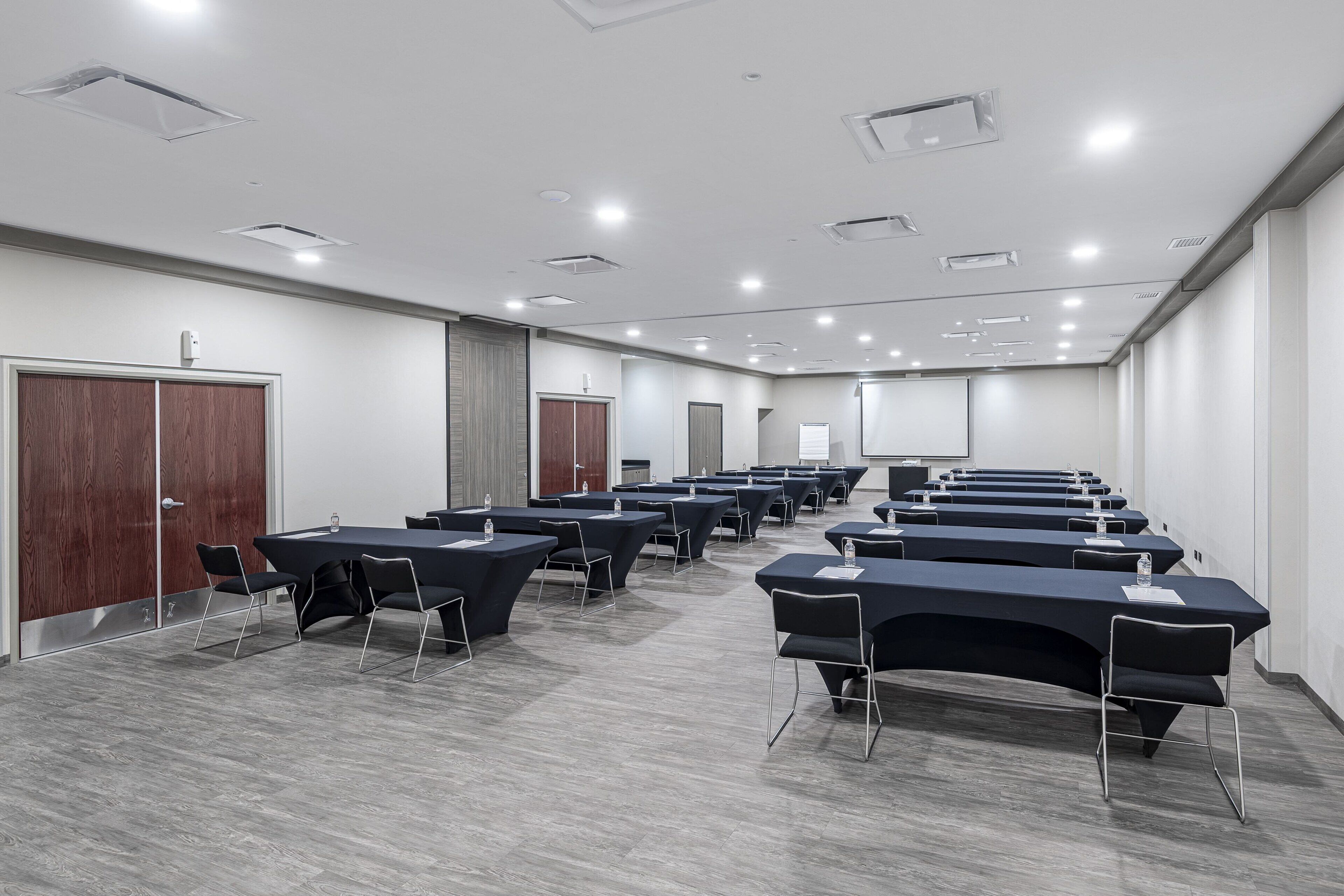 A conference room with multiple tables and chairs, wooden doors, and a white ceiling with recessed lighting.