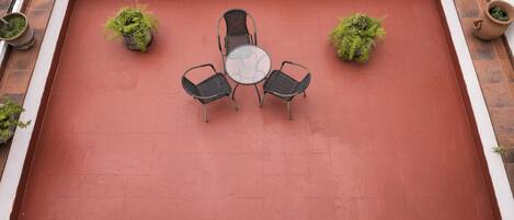 A rooftop patio with a red floor, surrounded by potted plants, and featuring a glass-top table with four chairs.