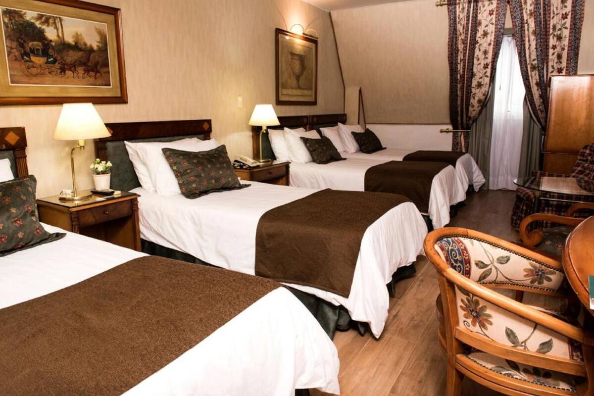 A hotel room with three beds, each with a brown blanket and white linens. There are two bedside tables with lamps, a wooden chair with floral upholstery, and a painting on the wall.
