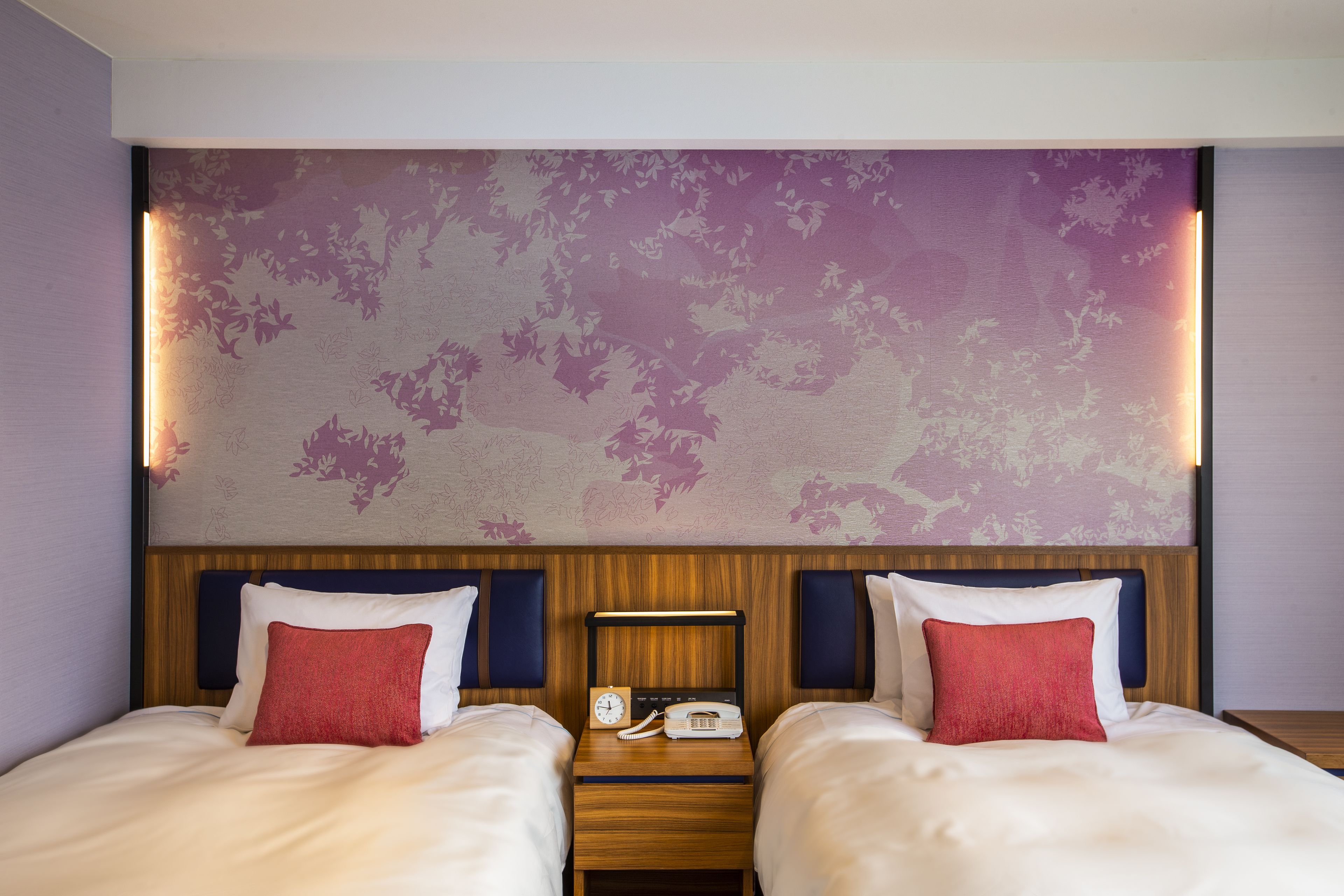 Een hotelkamer met twee bedden, een houten hoofdeinde, een nachtkastje met een klok en telefoon, en bloemenbehang in paarse tinten.
