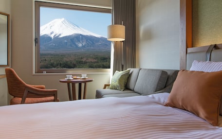 A hotel room with a bed, a sofa, a chair, a small table with a cup and saucer, and a view of a snow-capped mountain through a large window.